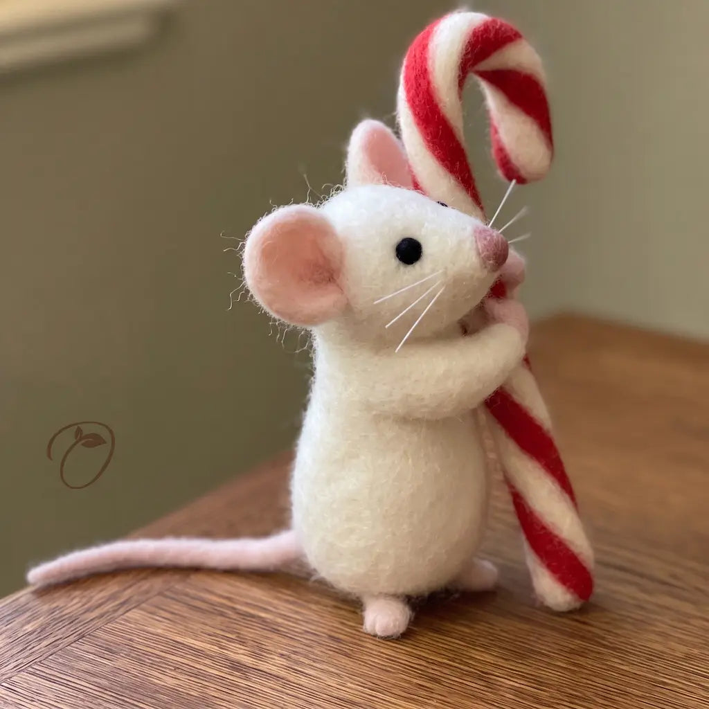 Side view of a wool felt mouse holding a striped candy cane, showing its pink nose, whiskers and long tail.