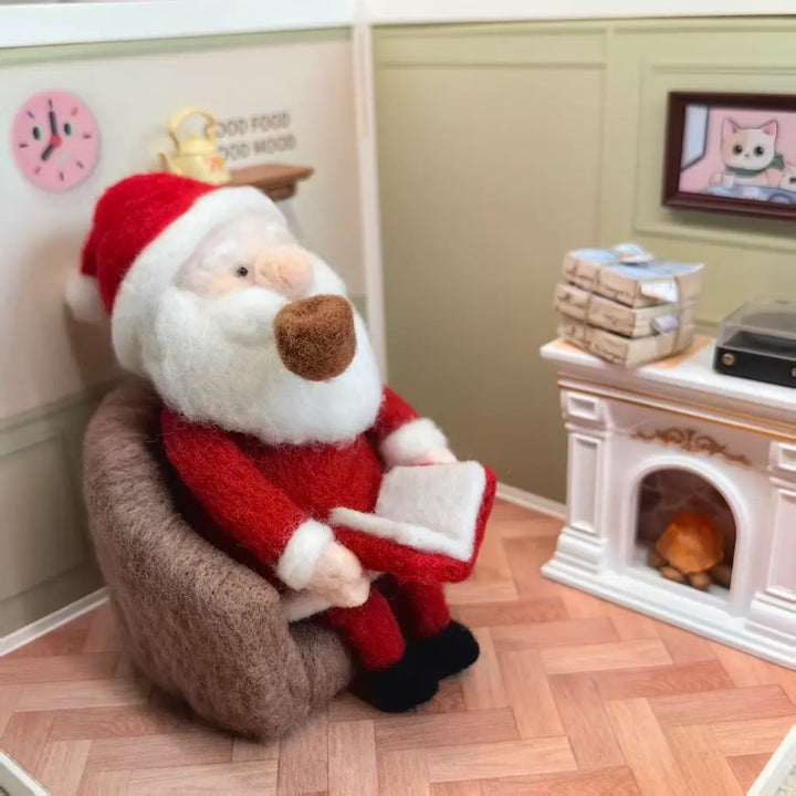 A felt Christmas ornament Santa Claus sitting in a brown armchair and reading a book, set inside a miniature dollhouse scene with a fireplace, wrapped gifts, and a wood-look floor.