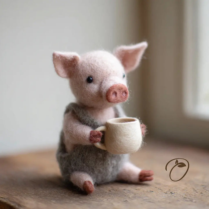 Three-quarter view of a seated pink wool felt piglet with a rosy snout, gray overalls, and a large cream mug.