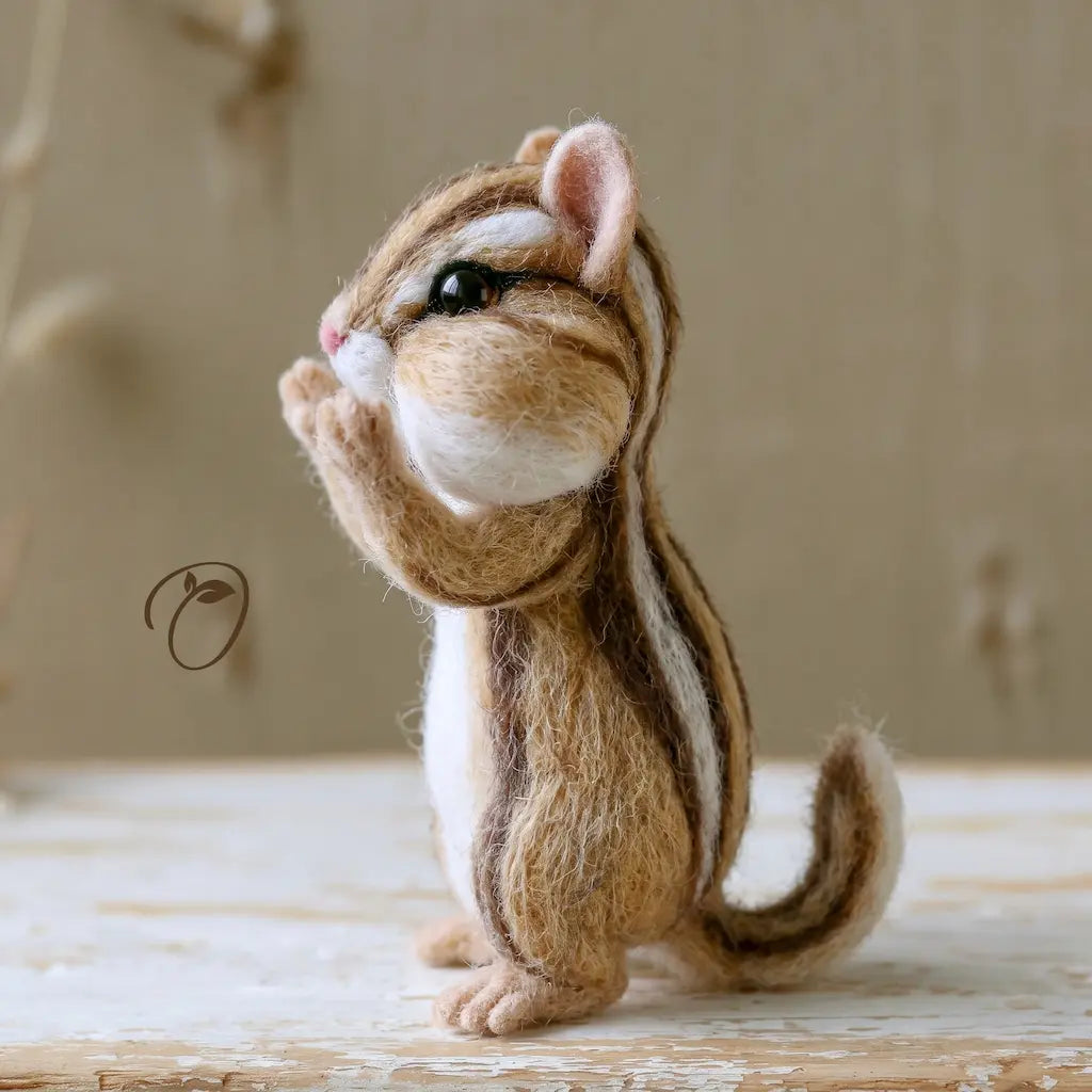 A needle-felted chipmunk figurine sitting upright on a rustic wooden surface against a soft beige background.