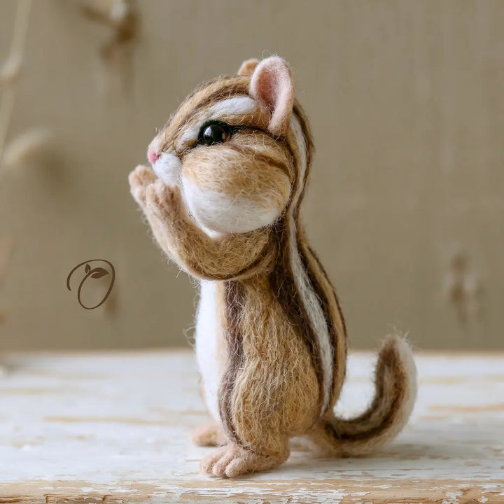 A needle-felted chipmunk figurine sitting upright on a rustic wooden surface against a soft beige background.