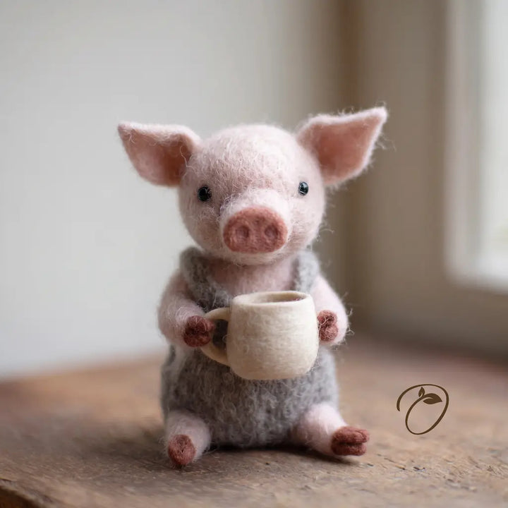 Front view of a pink felt piglet in gray overalls, holding a cream-colored mug in both front hooves.