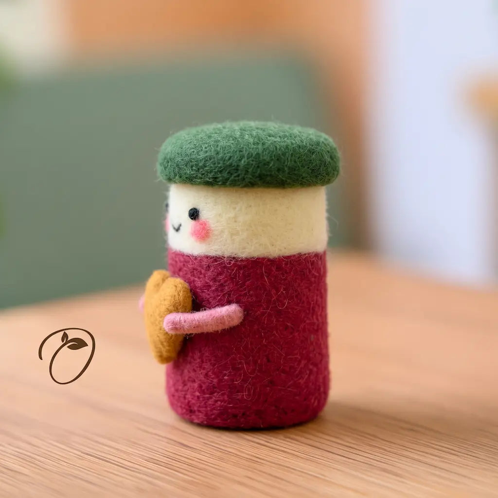 Side view of a cranberry jar felt figurine showing its rounded green lid, curved pink arm, and raised golden heart.