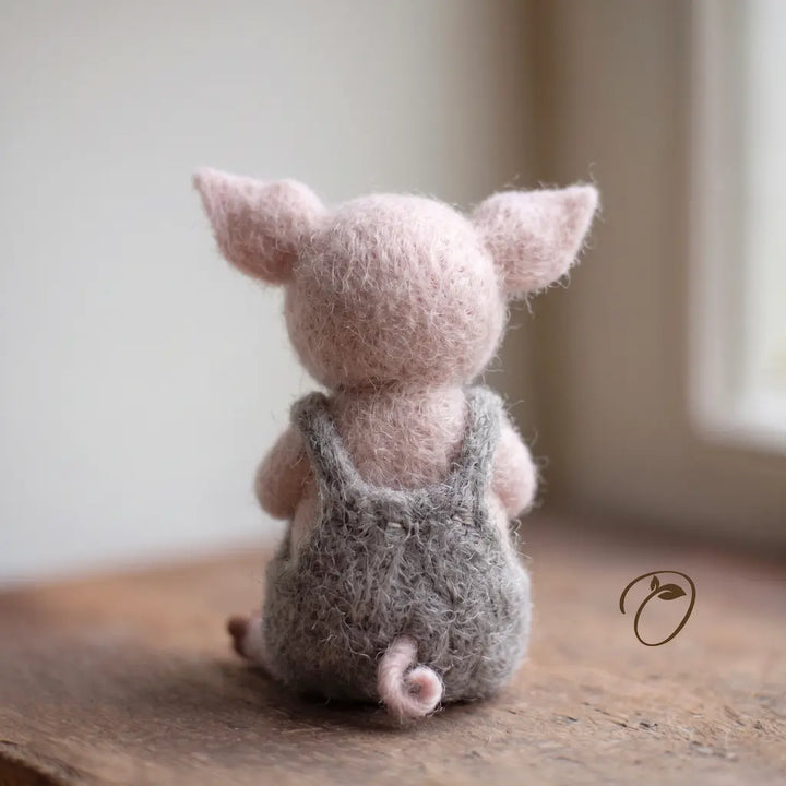 Back view of a pink felt piglet, showing gray overall straps, small stitching details, and a curled pink tail.