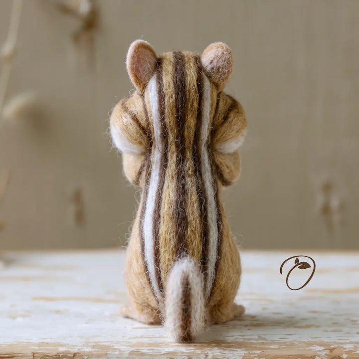 A needle-felted chipmunk figurine shown from the back, crafted with brown, beige, and white stripes, sitting on a rustic white wooden surface against a soft beige background.