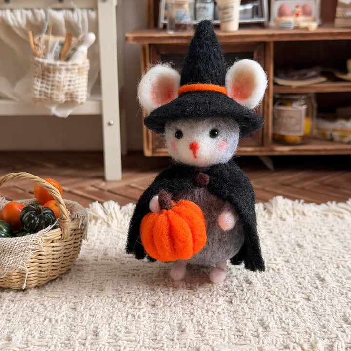 A small felt mouse doll dressed as a witch, wearing a black pointed hat with an orange band and a black cape, holding an orange pumpkin. The doll is standing on a cream rug in a cozy room setting with a basket of miniature pumpkins nearby.