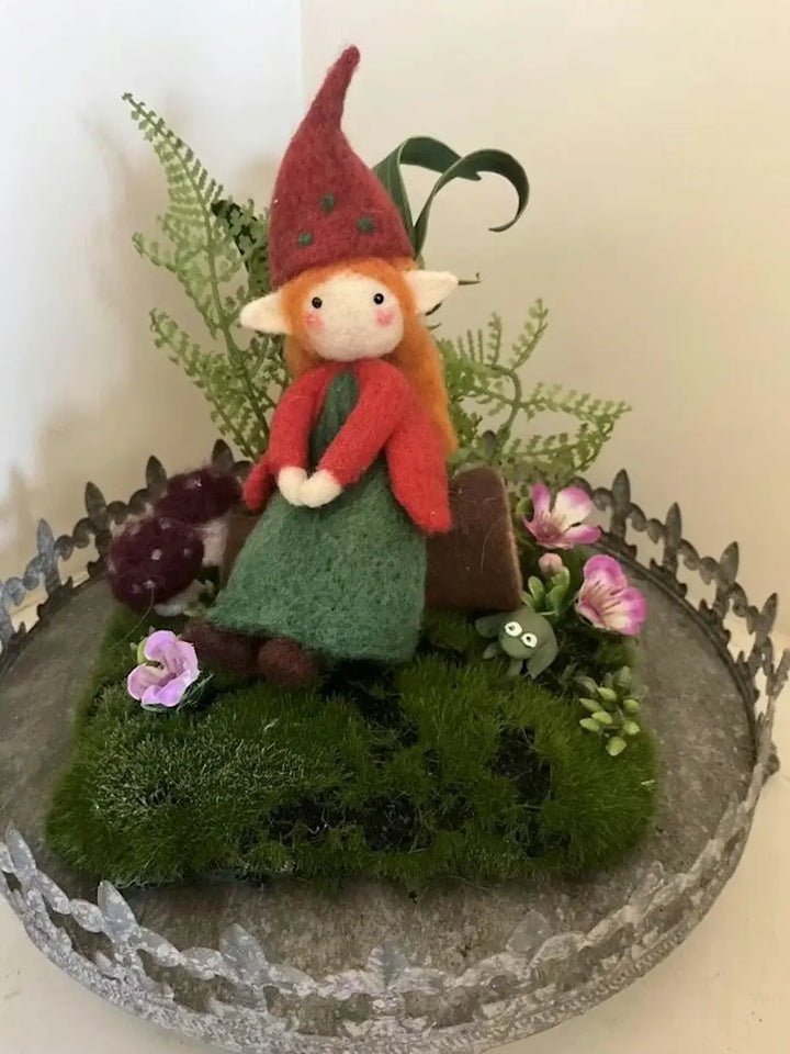 Elf figurine with red hat and green dress on a decorative stand with plants and flowers.