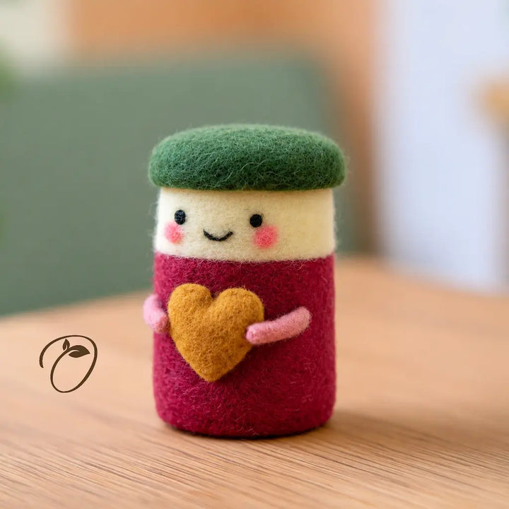 Front view of a cranberry-red wool felt jar with a green lid, smiling cream face, and pink arms holding a golden heart.