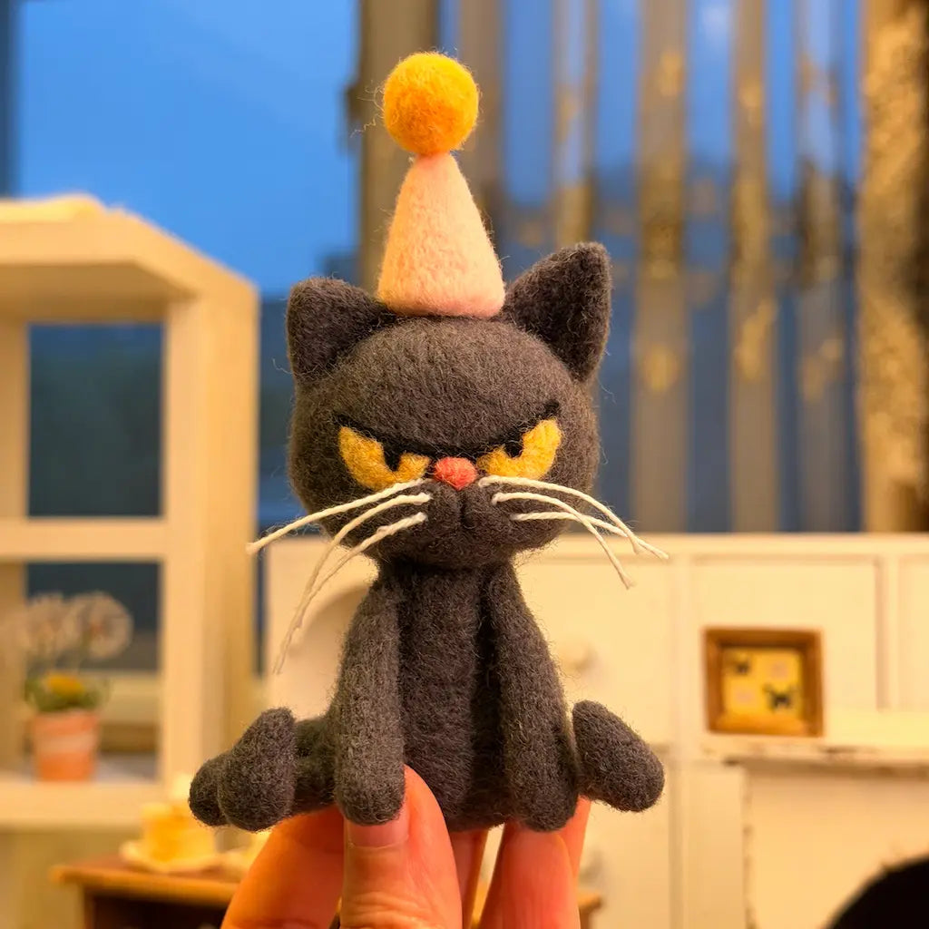 Front view of Birthday Mood Cat felt figurine showing its narrow golden-yellow eyes, pink nose, long white whiskers, and pink party hat with a yellow pom-pom.