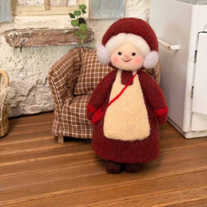 Handmade Burgundy Bonnet Mrs. Claus felt figurine wearing a burgundy dress, cream apron, and red crossbody bag, displayed beside a cozy plaid sofa.