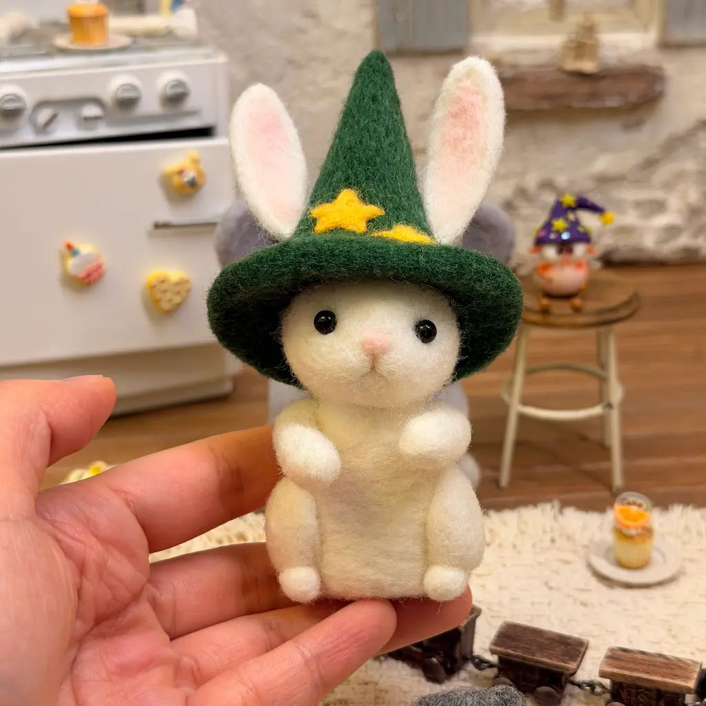 Hand-held front view of Starlit Cauldron Bunny felt figurine, featuring a white wool bunny with pink ears, glossy black eyes, and a green wizard hat decorated with yellow stars.