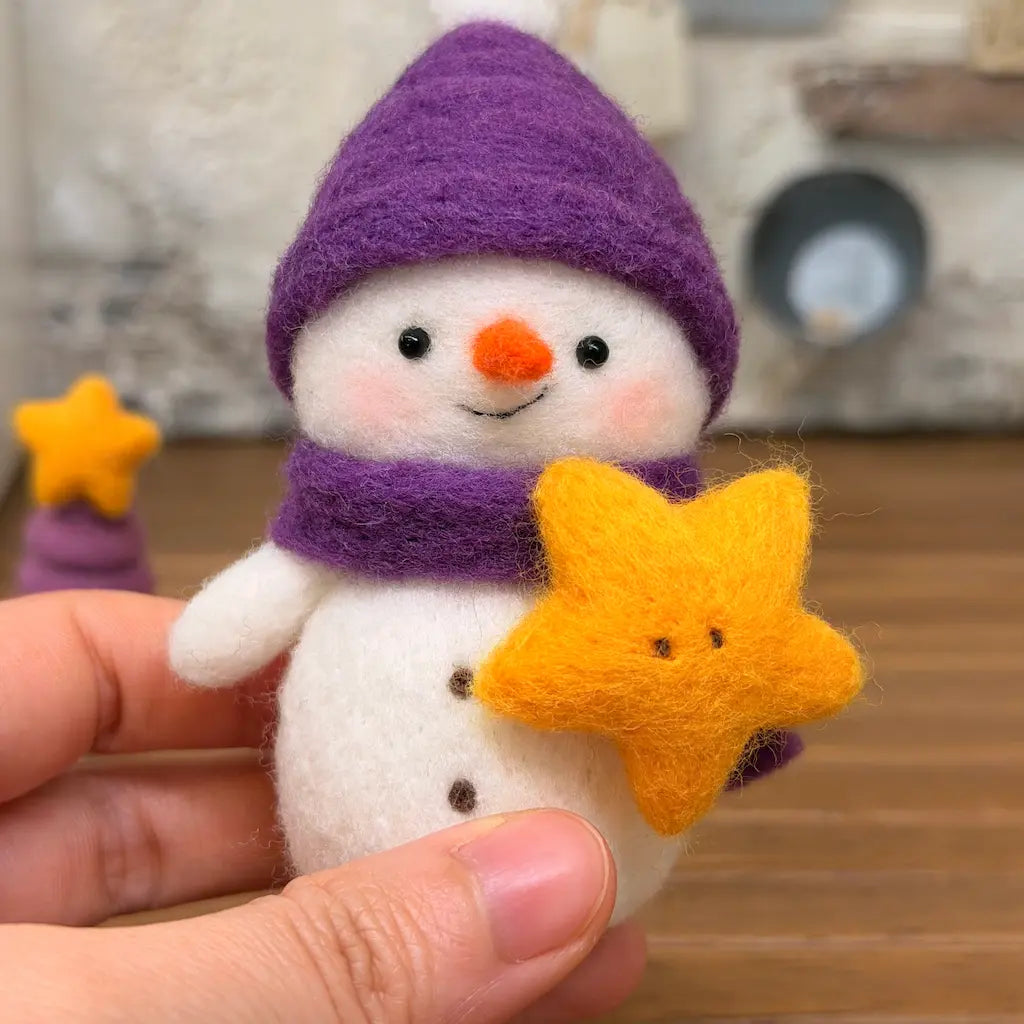 Hand-held close-up of Starlight Snowman wool felt figurine wearing a purple pom-pom hat and matching scarf, holding a smiling golden-yellow star.