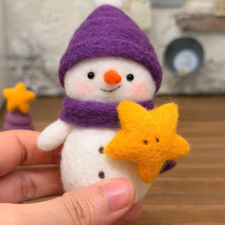 Hand-held close-up of Starlight Snowman wool felt figurine wearing a purple pom-pom hat and matching scarf, holding a smiling golden-yellow star.