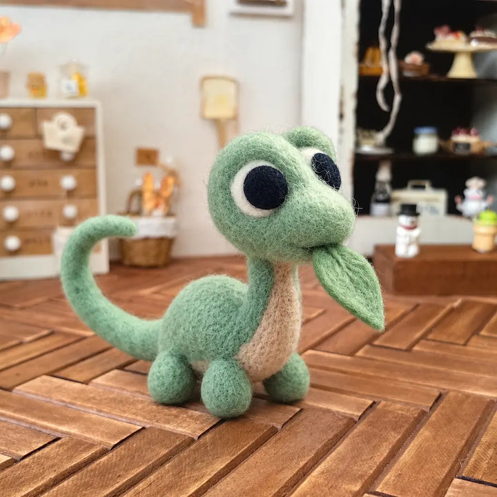Mint green wool felt dinosaur holding a leaf in its mouth on a wooden tabletop, with miniature shelves in the background.