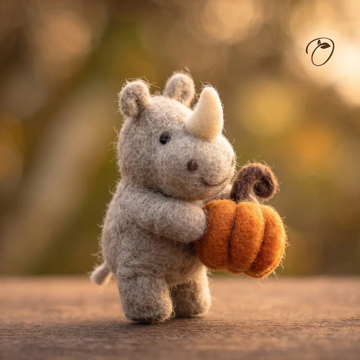 Three-quarter view of a gray felt rhino holding an orange pumpkin, showing its rounded muzzle, cream horn and short tail.