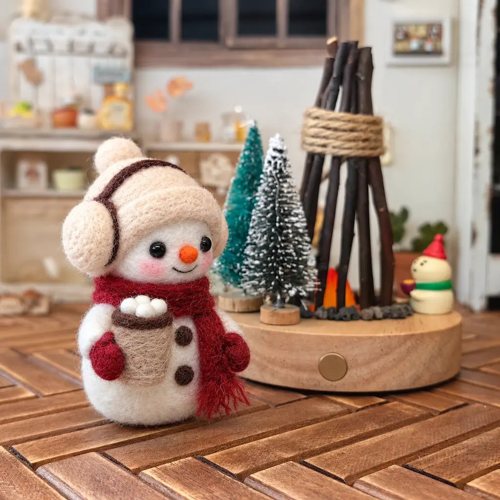 Handmade wool felt snowman figurine holding a tiny cocoa cup, wearing earmuffs and a red scarf, displayed on a wooden table with miniature Christmas trees and a cozy winter home background.