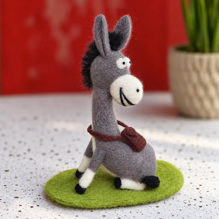 Three-quarter view of a gray felt donkey with a broad smile and brown satchel on a green mat against a red background.