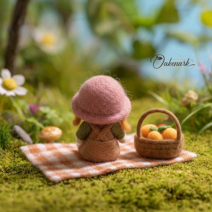 Small figurine of a mushroom with a pink cap and green body, sitting on a checkered cloth with a basket of oranges in a garden setting.
