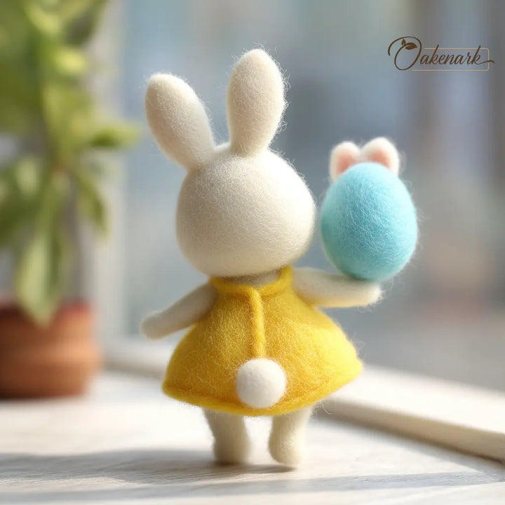 Small rabbit figurine in a yellow dress holding a blue egg, with a blurred plant and light background.