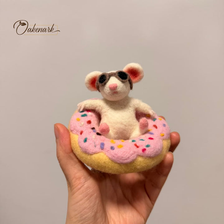 Felted mouse toy on a pink donut with sprinkles held in a hand against a plain background.