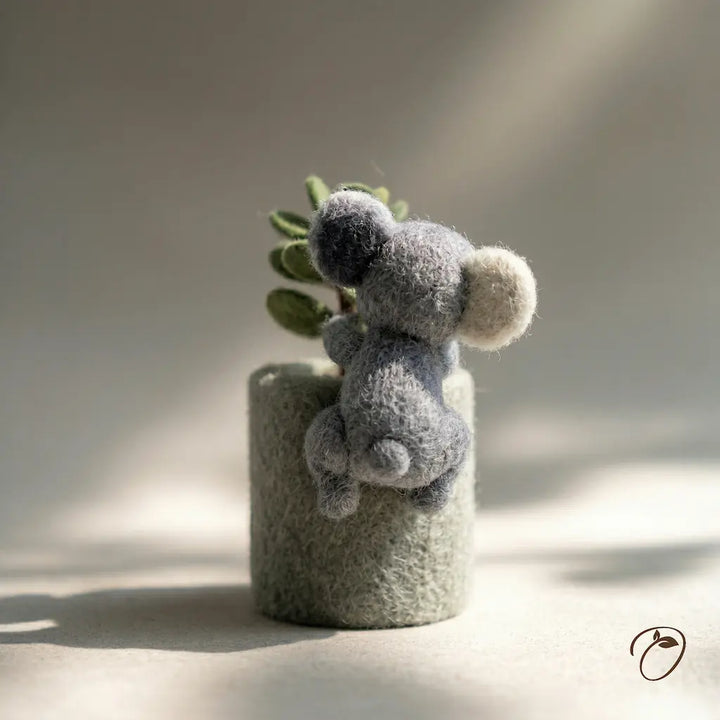 Gray fuzzy toy resembling a bear on a cylindrical gray base with a plant, against a neutral background.