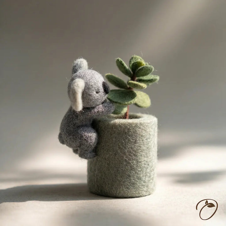 Small gray elephant plush toy holding a green plant in a textured pot on a neutral background