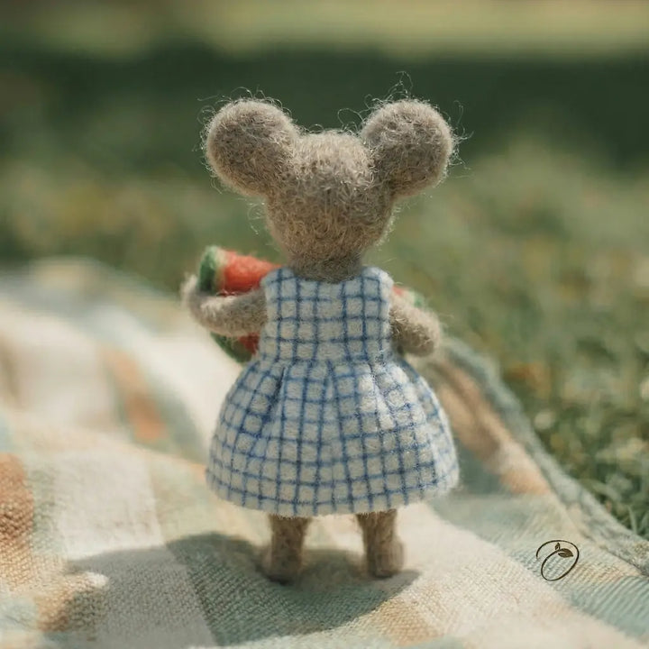 Small mouse figurine in a blue checkered dress holding a carrot, standing on a textured surface with a blurred natural background.