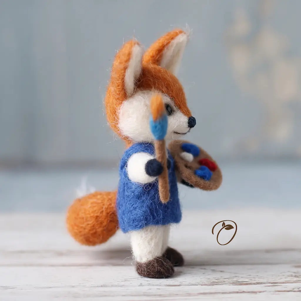 Felted fox figurine in a blue coat holding a paintbrush and palette on a wooden surface.