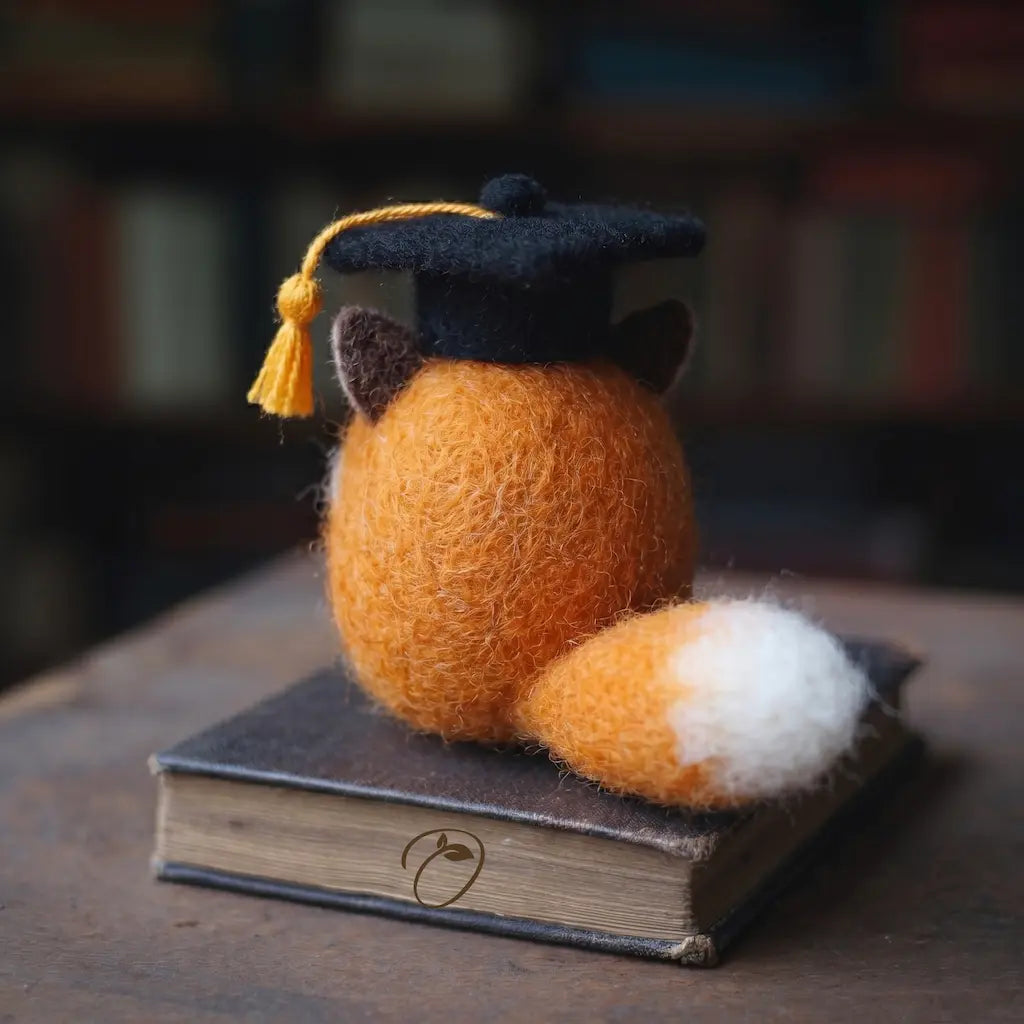 Felted fox figurine wearing a graduation cap on a book with a blurred bookshelf background