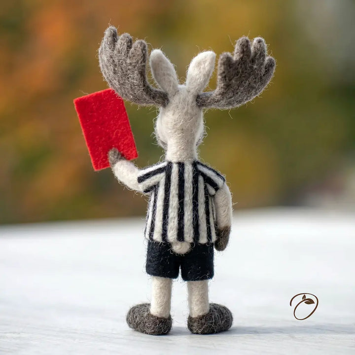 Toy moose in a referee outfit holding a red card against a blurred natural background