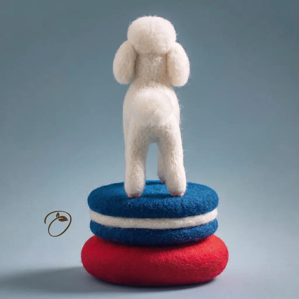 Felted toy sheep standing on a stack of red and blue felted objects against a gray background