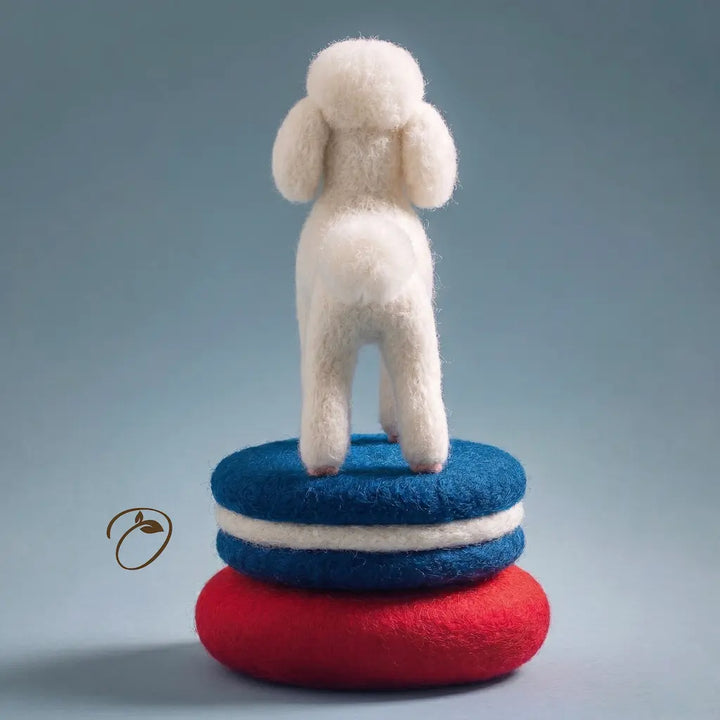 Felted toy sheep standing on a stack of red and blue felted objects against a gray background