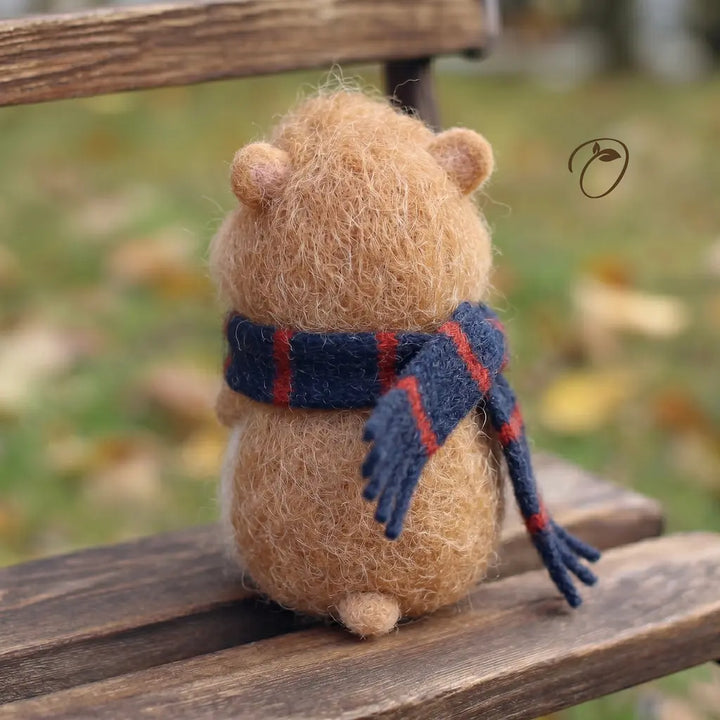 Small brown plush bear with a blue scarf sitting on a wooden bench in an outdoor setting.