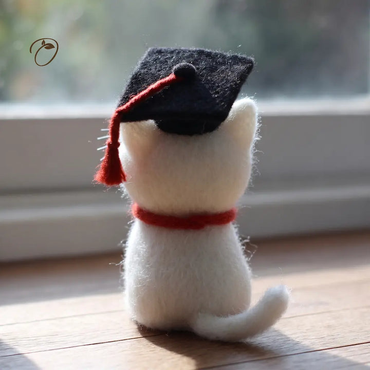 White cat-shaped plush toy wearing a black graduation cap and red tassel on a wooden surface.