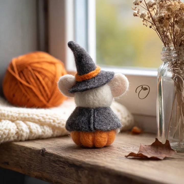 Small pumpkin figurine with a witch hat on a wooden surface next to yarn and a vase.