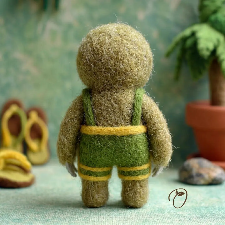 Small felted figure wearing green overalls with a blurred natural background