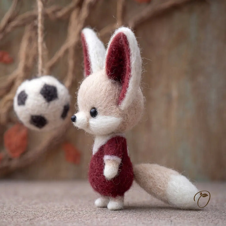 Felted fox toy with a red outfit standing on a textured surface with a blurred background.