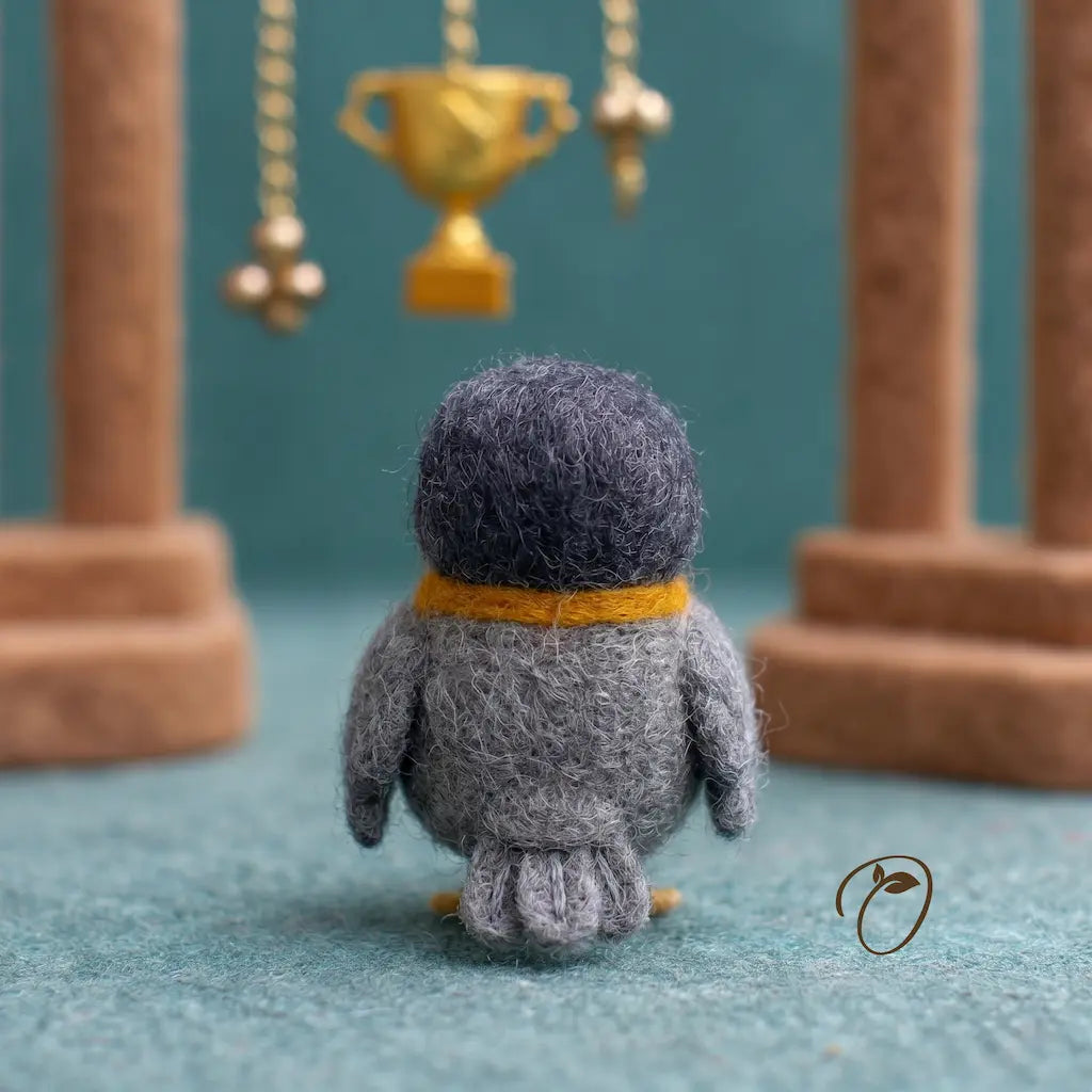 Small gray bird figurine with a yellow scarf standing on a blue surface, facing a golden trophy.