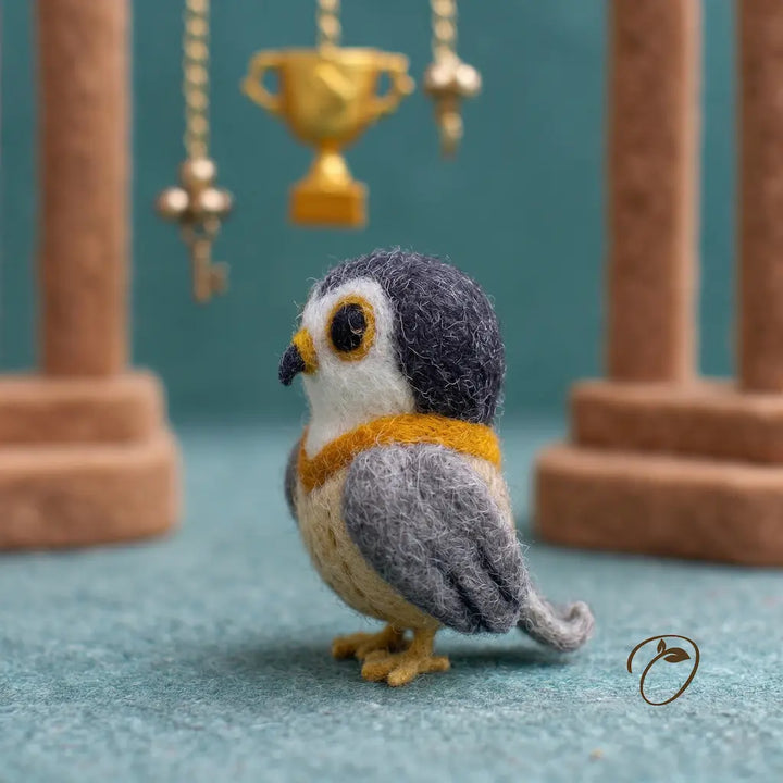 Felted owl figurine with a gold necklace on a teal surface with decorative columns in the background.