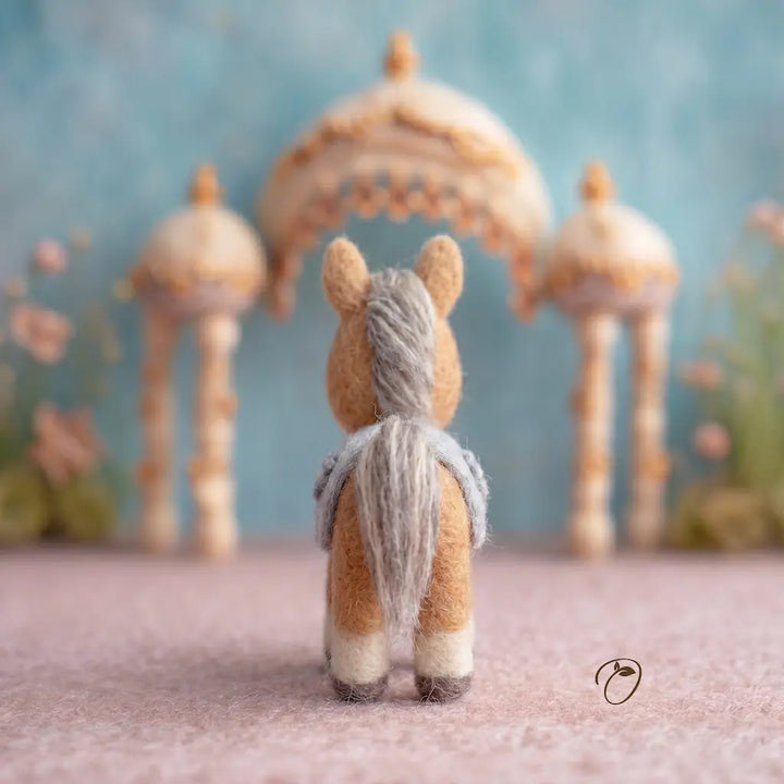 Small stuffed animal with a yarn tail in front of decorative arches and flowers.