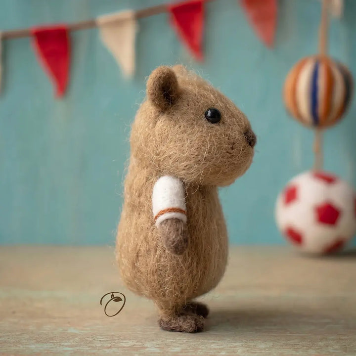 Felted animal toy resembling a beaver with a bandaged paw, standing on a wooden surface with a colorful background.