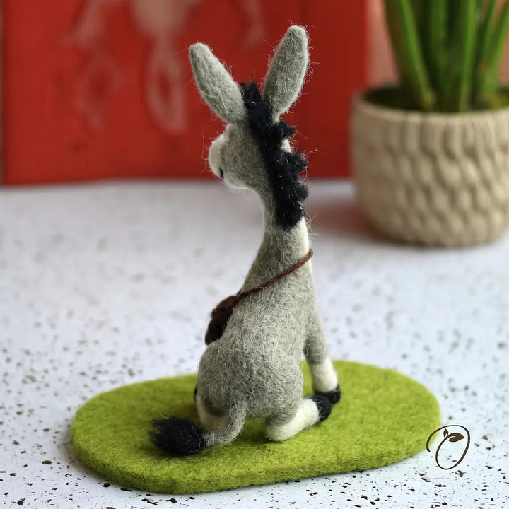 Felted donkey figurine on a green base with a blurred red object and plant in the background.