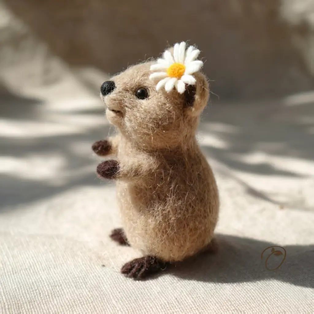 Small felted animal figurine with a white flower on its head, sitting on a textured surface.