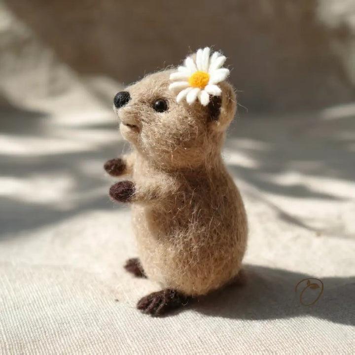 Small felted animal figurine with a white flower on its head, sitting on a textured surface.