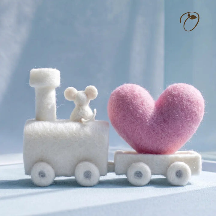 A small felt toy train with a white locomotive and carriages carrying a pink heart-shaped passenger, photographed against a soft light blue background.