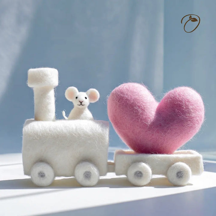 A small felted wool figurine train with a white engine and a pink heart-shaped carriage, featuring a tiny white mouse figure, set against a soft light blue background.