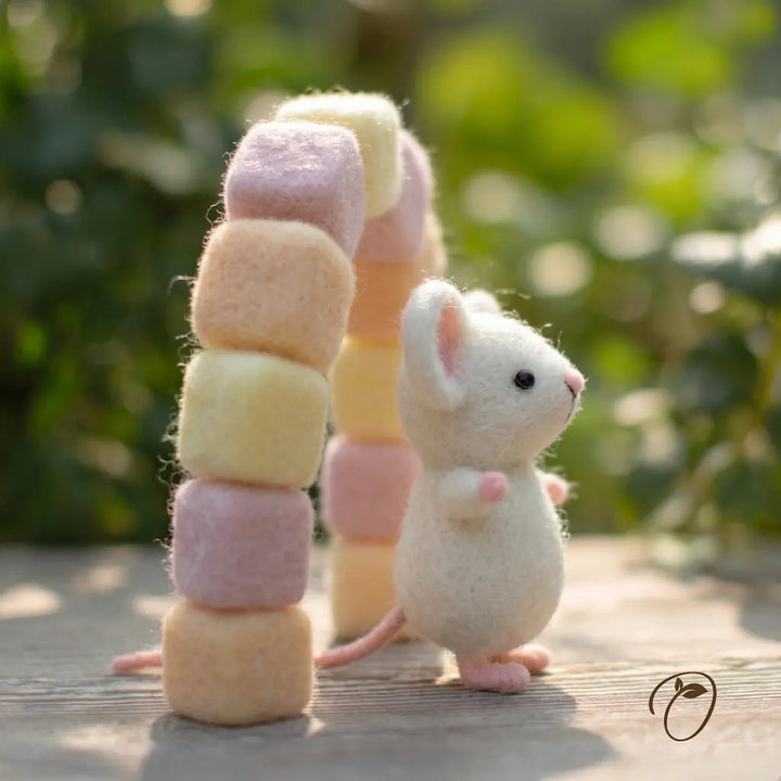 A small white felt mouse stands beside a pastel felt arch made of stacked mushroom shapes in pink, cream, and pale yellow, set outdoors on a wooden surface with a blurred green garden background.