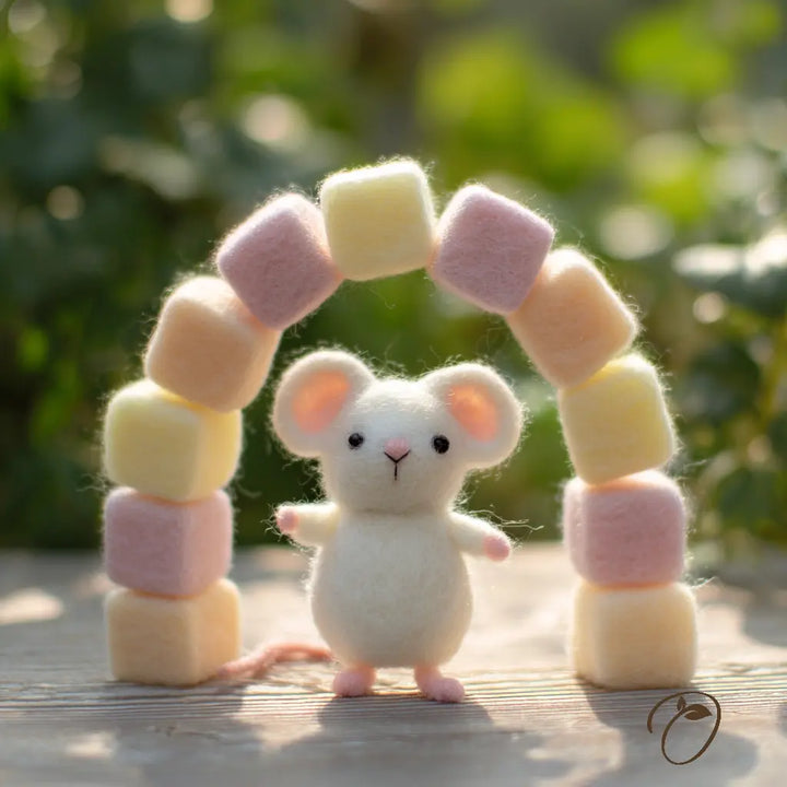 A small white felt mouse stands in front of a pastel felt arch made of square blocks in pale pink, cream, and peach, photographed outdoors on a wooden surface with blurred green foliage in the background.