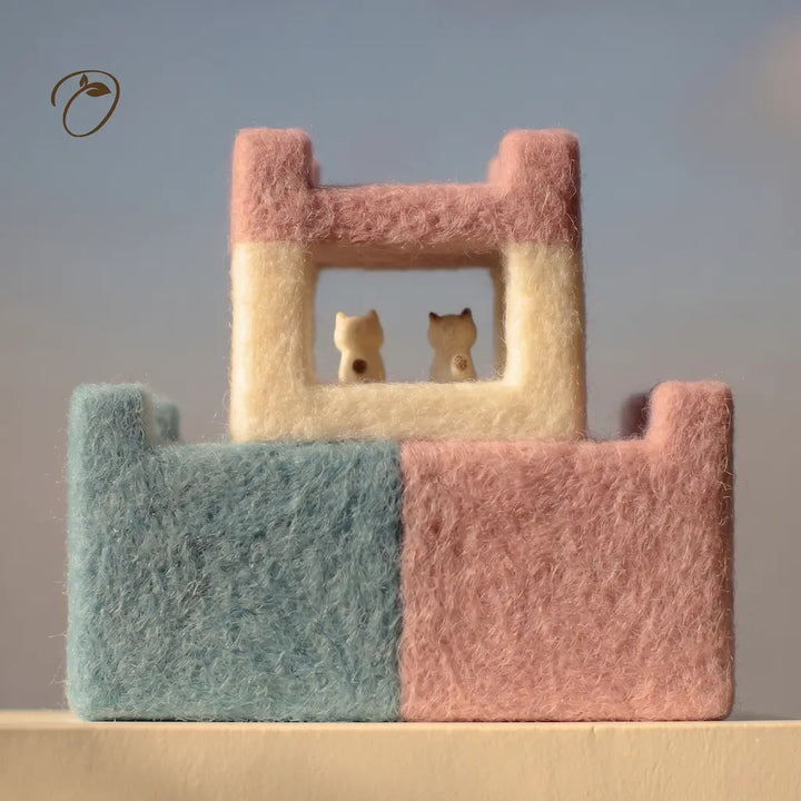 A pastel felt decorative house with a light blue base, pink upper walls, and a cream window frame, featuring two small cream cat figurines inside. The product is photographed on a plain surface against a soft blue-gray background.