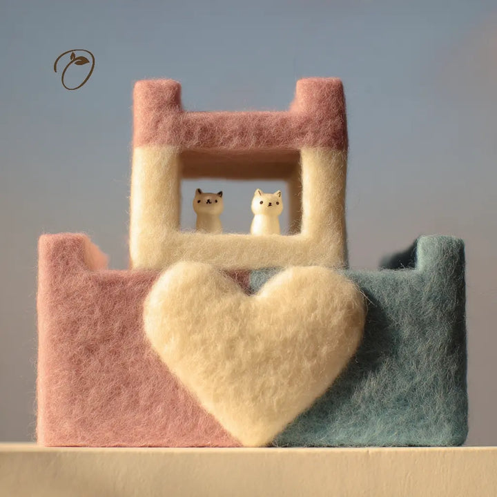 A pastel felt decorative structure resembling a small house or shelf, with a large heart-shaped cushion in front. The top section is cream with a pink roof, and the lower left section is pink while the lower right is muted blue-green.